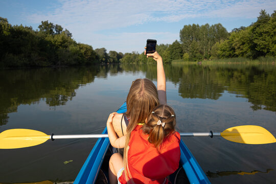 Mother taking a selfie with her daughter while kayaking on a calm lake surrounded by green trees on a sunny summer day. Happy family outdoor recreation and nature adventure