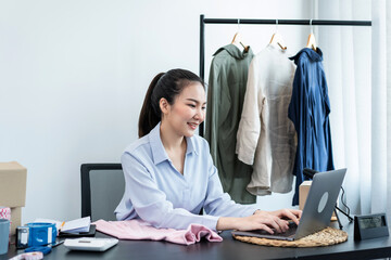 Female fashion designer is checking order customer on laptop and preparing packing clothes in cardboard boxes