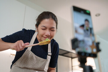 Asian woman in apron enjoys tasting food while filming cooking video with smartphone at home kitchen for social media.