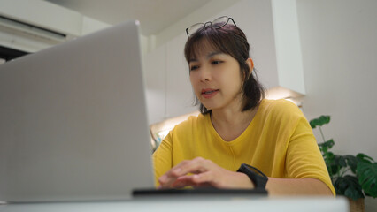 Focused Asian woman wearing yellow shirt typing on laptop at home, working remotely and managing...