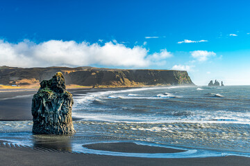 Basalt sea stack rises from the black sands of Reynisfjara Beach near Vík í Mýrdal, Iceland, with Atlantic waves and Reynisdrangar sea stacks in the distance.