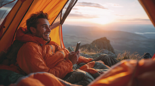 Smiling man relaxes in tent on mountain at sunrise, wearing warm gear and using smartphone, capturing a joyful outdoor adventure moment in nature.