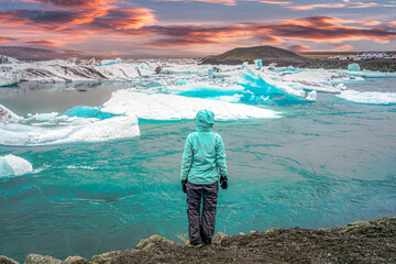 A traveler in a turquoise jacket watches floating icebergs at Jokulsarlon glacier lagoon in Iceland during a colorful sunset, symbolizing adventure and solitude. © LeeSensei