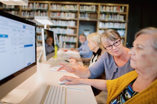 Elderly women using computers in a library. Elderly women learning technology in a computer class. Elderly women receiving computer assistance in a class. Elderly women in a library setting.
