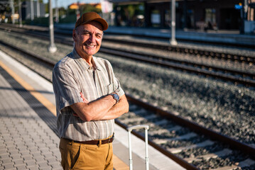 Fototapeta premium Happy senior man is standing at railway station and waiting for arrival of the train. Senior people traveling.