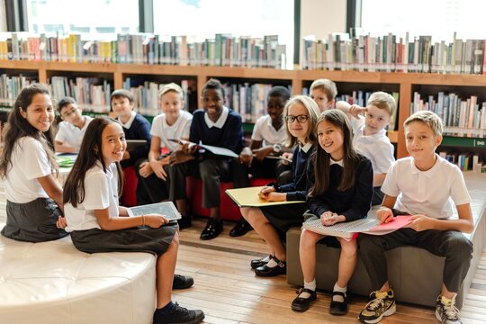 Group of diverse children in school uniforms sitting in a library, smiling and holding books. Children, library, and books create a learning atmosphere. Diverse young kids people in library.