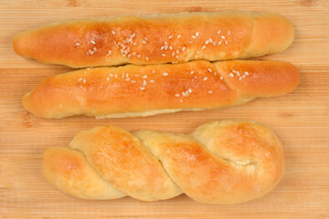 An overhead shot displays three freshly baked, golden-brown savory rolls - two sprinkled with sesame seeds and one braided - on a light wooden cutting board