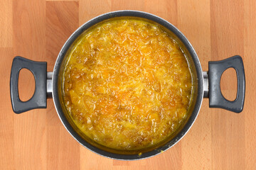 An overhead shot captures a thick, vibrant orange mandarin marmalade bubbling vigorously in a metal saucepan with gray plastic handles, resting on a wooden cutting board