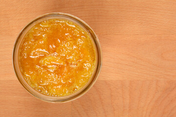 An overhead shot captures a clear glass jar filled with vibrant, chunky mandarin marmalade with visible pieces of peel, set on a light wooden surface with ample copy space