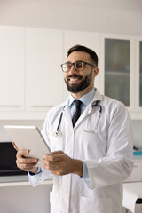 Fototapeta premium Smiling male doctor stands confidently in medical office, holding tablet