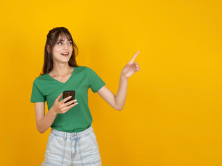 Woman pointing copy space, young caucasian 20s brunette woman pointing copy space with finger hold use mobile phone app looking workspace area isolated on yellow studio background. Design element idea