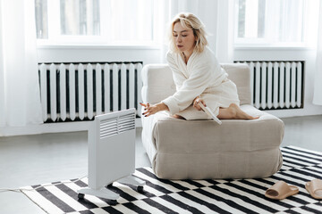 Young woman in a robe warming hands near electric heating radiator indoors