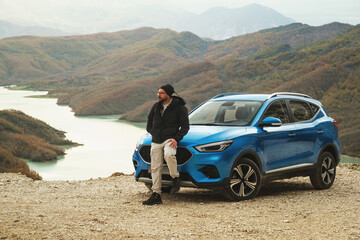 Blue colored suv, captured from a front angle emphasizing its left side, with Bovilla Lake in the background, accompanied by a man in a black jacket posing beside it