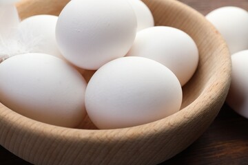 Raw chicken eggs in bowl and feather on wooden table, closeup