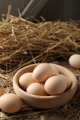 Raw chicken eggs in bowl and straw on wooden table, closeup