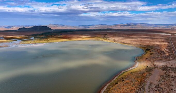 Flying over the lake with unclear water in the vast beautiful desert. Mountains with no vegetation at backdrop. Cumulus cloudscape covering the sky. Aerial view.