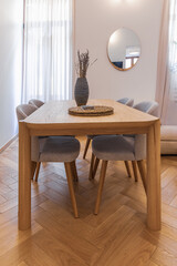 Close-up of a wooden dining table and fabric chairs in a small apartment interior. Features herringbone parquet floor and lavender decor in neutral tones
