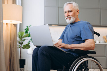 Senior man sitting in wheelchair working on laptop at home. Mature male browsing
