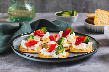 Appetizer of crackers with ricotta, cucumber and tomato on a plate on a table