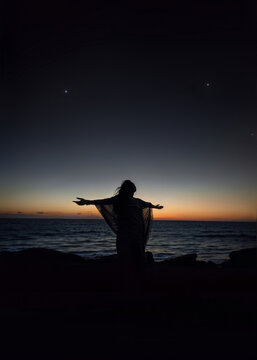 Silhouette of a woman standing on the beach at sunset with open arms toward the ocean, symbolizing freedom, peace, and mindfulness.