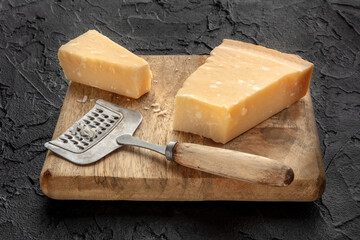 Parmesan cheese wedge on a wooden cutting board, with a cheese grater, on a black slate background