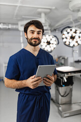 A surgeon stands in an operating room, holding a tablet and smiling at the camera. © sofiko14