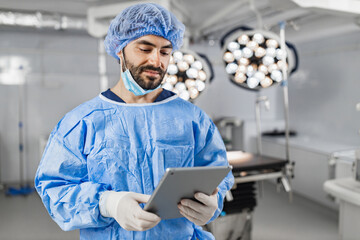A surgeon in sterile attire reviews a tablet in a modern operating room, with surgical lights and equipment in the background.