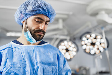 A surgeon wearing a surgical cap and mask in an operating room, looking down with a focused expression.