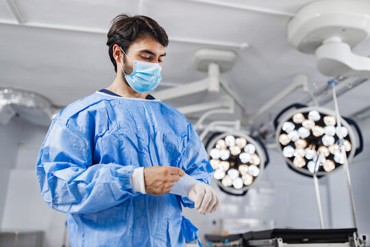 A surgeon in an operating room is putting on gloves, preparing for a medical procedure under bright surgical lights.
