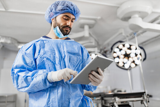 A surgeon in scrubs and a surgical cap uses a tablet in a modern operating room, likely reviewing patient information or surgical plans.