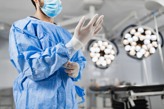 A surgeon in a blue surgical gown and mask puts on gloves in a bright operating room, preparing for a procedure.