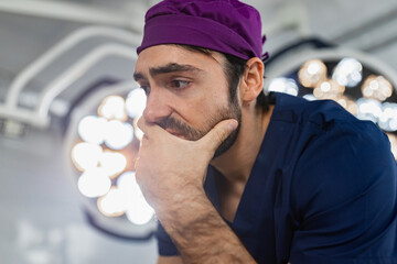 A surgeon in scrubs contemplates in an operating room, with surgical lights in the background.