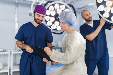 A surgeon reviews paperwork with a patient in an operating room, while another adjusts the surgical...