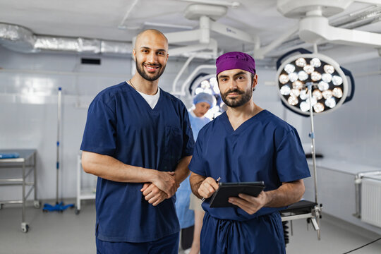 Two surgeons pose in an operating room, with surgical lights and equipment visible in the background, ready for a procedure. - Powered by Adobe