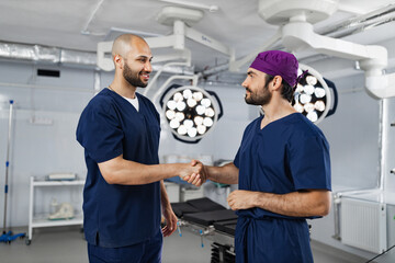 Two surgeons shake hands in an operating room, likely after a successful procedure or before a surgery. The room is well-lit with surgical lights.