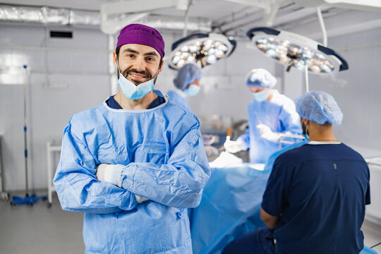 A smiling surgeon in scrubs stands in an operating room with his arms crossed, with other medical professionals in the background. - Powered by Adobe