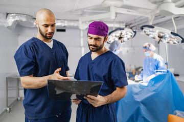 Two surgeons examine an X-ray while other medical professionals prepare for surgery in a sterile operating room.