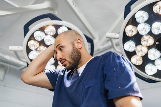A surgeon in a blue scrub suit looks concerned under bright operating room lights. - Powered by Adobe