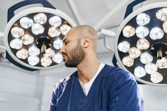 A surgeon stands in an operating room, illuminated by bright surgical lights, ready to perform a procedure.