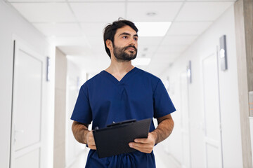 A doctor in blue scrubs walks down a clinic hallway, holding a clipboard. The image captures a...