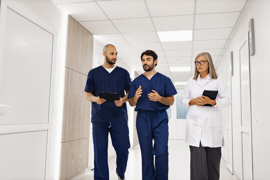 Three medical professionals, including a doctor, walk and discuss in a bright clinic hallway, holding tablets.