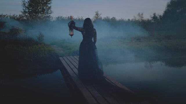 unrecognizable silhouette gothic woman hand hold lantern lamp walking across bridge over the river dark dense fog mist night nature mystical way. long black dress back rear view. Fantasy scared girl 