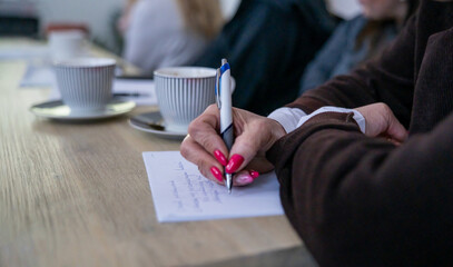 Close-up of a woman&rsquo;s hand writing notes during a meeting or workshop. Coffee cups and blurred participants in the background. Concept of learning, education, and creative process.