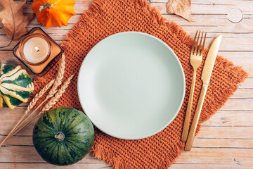 Autumn table setting with pumpkins, candle, plate, and golden cutlery on a brown napkin on wooden background. Cozy fall decoration. Top view, flat lay, mockup
