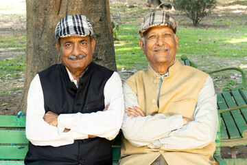 Indian happy two senior male friends arm crossed while spending leisure time at park. Old friends wearing jacket and golf cap. Happy elderly people enjoying life after retirement.