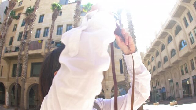 A woman takes a photo. A happy tourist. A brunette in stylish clothes poses with a camera in front of an Arab city. Adventures and travel.