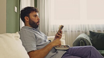 A bearded man is comfortably seated on a sofa, enjoying a snack while engrossed in his mobile phone. Experience the authentic sounds of him munching on cookies, adding to the relaxed atmosphere.
