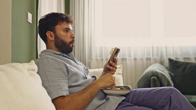 A bearded man is comfortably seated on a sofa, enjoying a snack while engrossed in his mobile phone. Experience the authentic sounds of him munching on cookies, adding to the relaxed atmosphere.