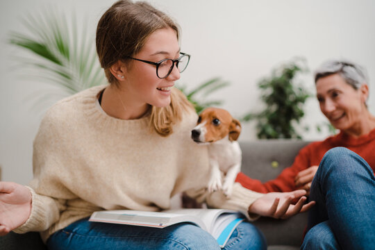 Young smiling woman in glasses sitting on sofa with book and playing with dog spending time with mature mother