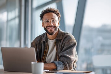 A happy man sitting at a table, using a laptop in a bright, modern home office. the man he is smiling while working online or looking for inspiration.